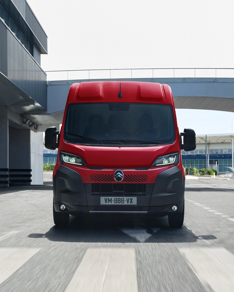 Vorderansicht eines roten Citroën Jumper, der durch eine moderne Stadtlandschaft fährt und dabei sein robustes Design, seine Zweckmäßigkeit und seine beeindruckende Straßenpräsenz unterstreicht.